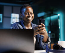 Cheerful female college student using mobile device to engage with friends on social media platforms. African american lady using cell phone to surf the web while sitting at desk in her home office.